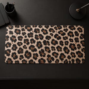 May include: A rectangular desk mat featuring a repeating leopard print pattern with black and dark brown spots set against a light tan background. The mat has rounded corners and a smooth, flat surface, providing a consistent texture throughout.