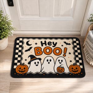 Puede incluir: Un felpudo rectangular con un centro color crema y bordes a cuadros negros. El texto 'Hey BOO!' está impreso en negro y naranja. El diseño presenta tres fantasmas blancos, dos calabazas naranjas, una luna creciente y murciélagos.