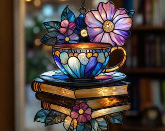 May include: Stained glass suncatcher featuring a teacup, saucer, and books. The teacup is blue with a gold handle, resting on a stack of books. Flowers in shades of purple, pink, and blue adorn the top and bottom. A decorative piece.