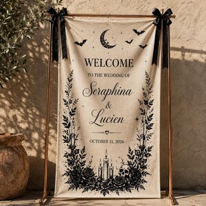May include: A rectangular beige fabric wedding welcome sign hanging from a copper-colored metal stand with black ribbon bows. The sign features black printed text reading Welcome to the wedding of Seraphina & Lucien October 31, 2026, with moon, bat, candle, rose, and skull illustrations.