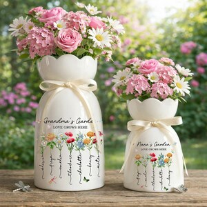 Puede incluir: Dos jarrones de cerámica blanca con bordes festoneados, llenos de flores rosas y blancas. El jarrón más grande dice "Grandma's Garde, Love Grows Here" con ilustraciones florales y nombres. El jarrón más pequeño dice "Nana's Garden, Love Grows Here".