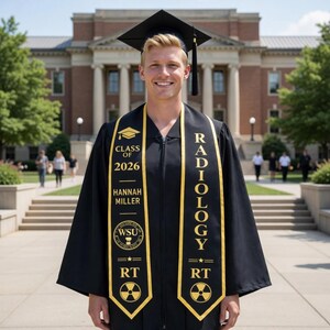 May include: A person in a black graduation gown and cap, smiling in front of a brick building. The gown has a yellow sash that reads "RADIOLOGY" and "RT". The sash also has the text "CLASS OF 2026" and "HANNAH MILLER".