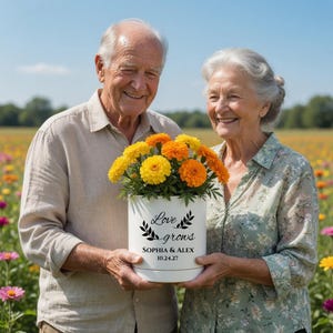 Może przedstawiać: Starsza para trzyma białą donicę z żółtymi i pomarańczowymi kwiatami. Na donicy widnieją słowa "Love grows", imiona "SOPHIA & ALEX" i data "10.24.27". Para uśmiecha się na polu kwiatów.