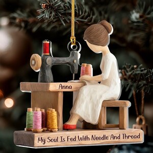 May include: A wooden Christmas ornament depicting a person sewing at a sewing machine. The ornament features the name "Anna" and the text "My Soul Is Fed With Needle And Thread". The ornament includes spools of thread in various colors.