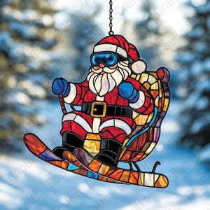 以下が含まれることがあります： ステンドグラスのクリスマスオーナメント。スキーをするサンタクロースが描かれています。サンタは赤いスーツ、帽子、青いゴーグルを着用しています。オーナメントは吊り下げ用の黒いチェーン付きで、雪景色を背景にしています。
