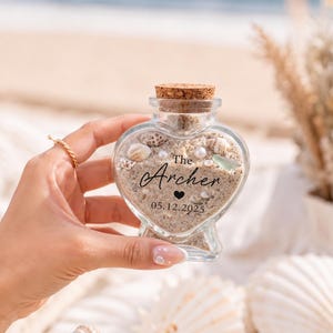 May include: A heart-shaped glass bottle with a cork stopper, filled with sand, small seashells, and white pearls. The front of the bottle features black printed text that reads The Archer, a small heart icon, and the date 05.12.2025.