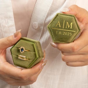Peut inclure: Deux boîtes à bagues hexagonales en velours vert olive, l'une ouverte avec deux bagues en or et l'autre fermée. La boîte fermée porte les initiales "A|M" et la date "1.6.2025" en lettres dorées. Les bagues ont une pierre précieuse bleue et de petits diamants.