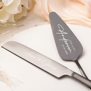 Könnte beinhalten: Ein silbernes Kuchenmesser- und Servier-Set. Der Servierer hat eine gezahnte Kante und den Namen "Anderson" sowie das Hochzeitsdatum "09-06-2025" eingraviert. Auf dem Messer stehen die Namen "Anna & James".