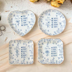 May include: Four decorative ceramic plates with floral patterns and gold trim. The plates are in various shapes: heart, round, square, and a scalloped square. Each plate features a blue floral design and text that reads "God Says I Am".