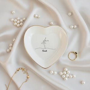 May include: A heart-shaped white ceramic trinket dish with a gold rim, featuring a minimalist ballerina illustration and the name "Sarah". The dish is surrounded by pearls, a gold ring, and a star necklace, all set on a white satin surface.