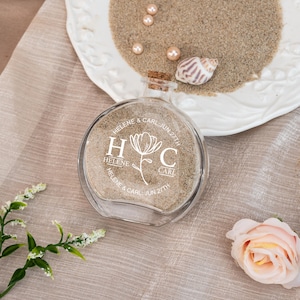 May include: A clear, round glass bottle filled with sand, sealed with a cork, and engraved with "Helene & Carl Jun 27th". The bottle features a floral design with the initials "H" and "C". A white plate with sand, pearls, and a seashell is in the background.