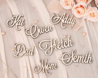 May include: Wooden name tags in a cursive font, including names like "Kai," "Addy," and "Owen." The tags are light brown and have small holes for hanging. The background includes fabric, pearls, and pink roses.