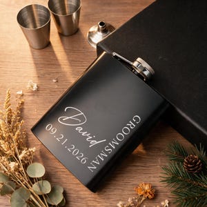 Op de afbeelding: Een matzwarte metalen heupfles met de gegraveerde naam David, de datum 09.21.2026 en het woord GROOMSMAN in witte letters. De heupfles heeft een zilveren schroefdop en ligt naast twee kleine zilveren metalen bekers en een zilveren trechter.