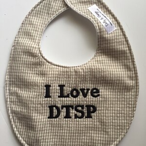 May include: A beige and white checkered baby bib with the embroidered text "I Love DTSP" in black. The bib has a snap closure and a small white tag with text.