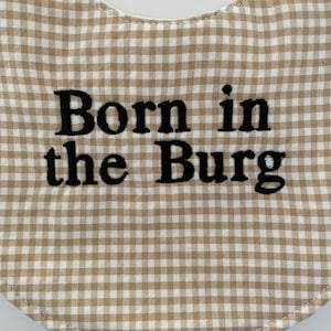 May include: A tan and white gingham baby bib with black embroidered text that reads "Born in the Burg".
