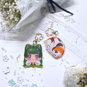 May include: Two acrylic charms with gold-colored hardware. One charm features a green-haired anime-style character with cat ears, and the other depicts a sleeping calico cat. Both charms have a floral accent.