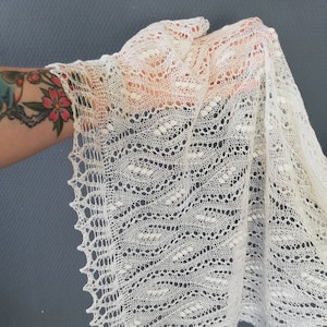 Puede incluir: Un chal de ganchillo blanco con un delicado patrón de encaje. El chal está drapeado sobre una mano y tiene un borde festoneado.