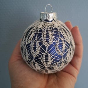 Op de afbeelding: Een blauwe glazen kerstbal bedekt met een wit gehaakt kantontwerp.