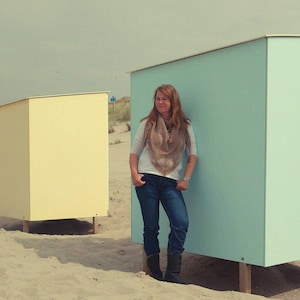 May include: A woman wearing a brown scarf and blue jeans stands in front of a light blue beach hut. A yellow beach hut is visible in the background.