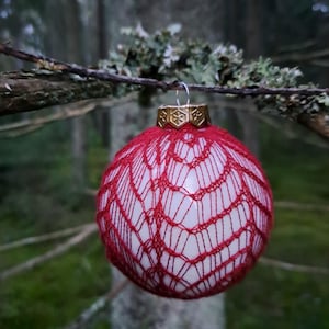 May include: A red and white Christmas ornament with a delicate, web-like design. The ornament has a gold-colored cap and is hanging from a tree branch. The background is a blurred forest scene.
