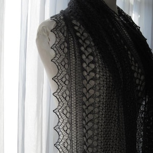 Op de afbeelding: Een zwarte kanten sjaal met een delicaat patroon, gedrapeerd over een paspop. De sjaal is gemaakt van fijn garen en heeft een zachte, vloeiende textuur.