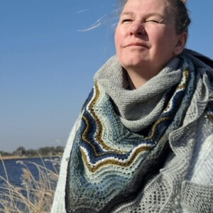 May include: A woman wearing a gray knitted sweater with a blue, green, and brown crocheted shawl. She is standing in a field of tall grass with a body of water in the background.