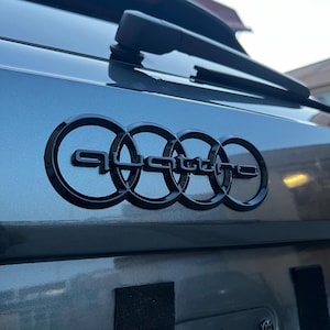 Puede incluir: Primer plano de la parte trasera de un coche gris con un emblema "Quattro" negro. El emblema está compuesto por cuatro anillos entrelazados con la palabra "Quattro" en una fuente estilizada. El limpiaparabrisas del coche es visible encima.