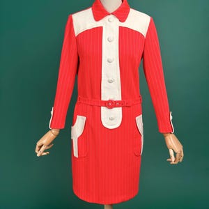 Puede incluir: Un vestido vintage rojo y blanco con rayas verticales. El vestido presenta un cuello blanco, una botonadura frontal y un cinturón a juego. Los detalles blancos están en los bolsillos y los puños. El vestido se exhibe en un maniquí.