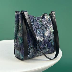 Peut inclure: Sac à main rectangulaire avec un motif peau de serpent dans les tons de bleu, vert et blanc. Le sac a des bordures et des poignées en cuir noir. Le sac est posé sur une surface blanche sur un fond vert.