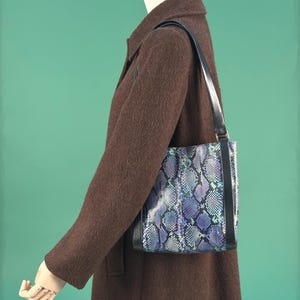 Könnte beinhalten: Ein dunkelbrauner Mantel mit einer schwarzen Umhängetasche. Die Tasche hat ein Schlangenmuster in Blau-, Lila- und Türkistönen mit schwarzen Zierleisten. Die Tasche hat eine rechteckige Form.