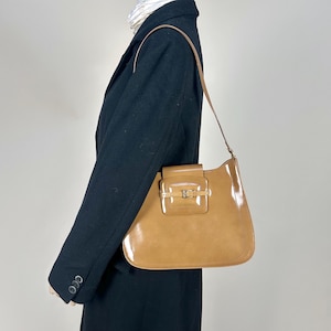 May include: A tan leather shoulder bag with a flap closure and a silver buckle. The bag has a shiny finish and a curved shape.
