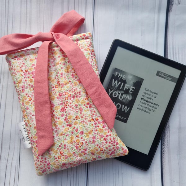 Könnte beinhalten: Eine pinkfarbene Buchhülle mit floralem Muster und einer rosa Schleife. Die Hülle hält einen schwarz-weißen E-Reader mit dem Text "The Wife You Know" auf dem Cover.