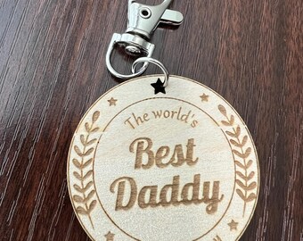 May include: A wooden keychain with a silver clasp. The round wooden pendant is engraved with the words "The world's Best Daddy" and the names "Bailey & Tommy". The design includes laurel leaves and star accents.
