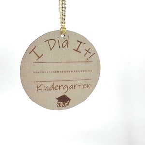 Puede incluir: Un adorno redondo de madera con una cinta dorada. Dice "¡Lo hice! Kindergarten 2025" con un icono de gorro de graduación.