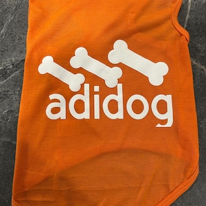 Puede incluir: Camiseta naranja brillante para perros con texto y gráficos de huesos blancos. La palabra "adidog" está impresa en letras blancas debajo de tres ilustraciones de huesos blancos. La camiseta está hecha de un material suave y ligero.