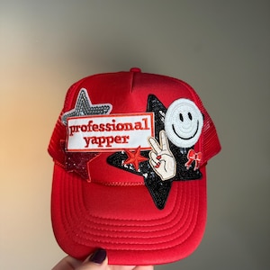 May include: A red trucker hat with a mesh back and a curved brim. The hat features the text "professional yapper" on a white rectangle, along with star and peace sign patches, and a smiley face.