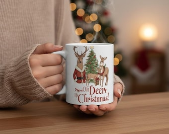May include: White ceramic mug featuring a festive deer family illustration with a Christmas tree and gifts. The deer are wearing Santa-style outfits. The mug has the text "Mr. Oh Deer It's Christmas!" in red script.
