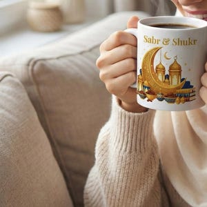 Peut inclure: Tasse en céramique blanche avec les mots "Sabr & Shukr" et un croissant de lune doré et un motif de mosquée. La tasse est remplie de café noir. La personne porte un pull crème.