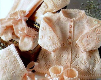 Könnte beinhalten: Ein Set Babykleidung in zartem Pfirsichton, bestehend aus Strickjacke, Mütze, Babyschuhen, Fäustlingen und einem Schal. Die Strickwaren weisen ein zartes Blättermuster auf und sind mit Satinbändern verziert.