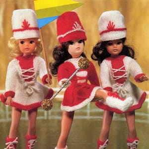 May include: Three dolls wearing red and white knitted outfits with matching hats. The dolls are holding a baton, a flag, and a drum.