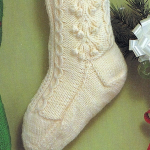 May include: A white knitted Christmas stocking with a cable knit pattern and a leafy design. The stocking is hanging on a green wall with a white bow and a candy cane.
