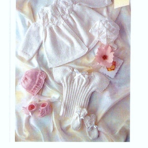 May include: A set of white knitted baby clothes, including a cardigan, a top, a hat, and booties. The clothes are laid out on a white satin fabric. The text "Premature Baby Attire" is visible in the top right corner of the image.