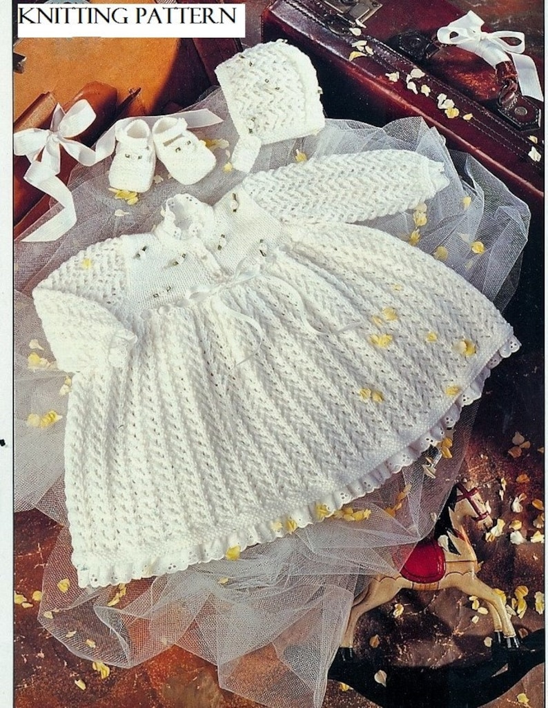 May include: A white knitted baby dress, bonnet, booties, and sleeves set. The dress has a textured pattern and a ruffled hem. The image also includes a white ribbon and the text "KNITTING PATTERN".