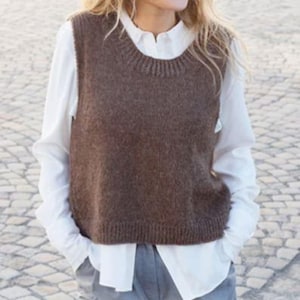 May include: A dark brown sleeveless knit sweater vest with a ribbed crew neckline and hem, layered over a crisp white long-sleeved button-down shirt. The vest features a textured wool-like knit construction and a straight, boxy fit.