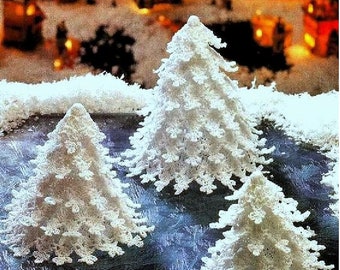 Puede incluir: Tres árboles de Navidad blancos, hechos a ganchillo, de diferentes tamaños. Los árboles están dispuestos sobre una superficie azul con efecto de nieve, y el fondo presenta luces borrosas, sugiriendo una escena invernal.