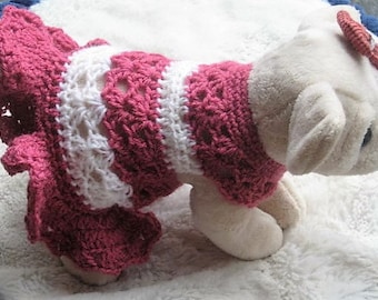 May include: A white stuffed dog wearing a pink and white crocheted dress with a pink flower on its head.