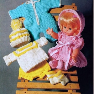 Könnte beinhalten: Ein Set gestrickter Puppenkleidung, bestehend aus einem blauen Overall, einem weißen Pullover mit gelben Streifen, einer gelben Hose, einer weißen Mütze mit gelben Streifen und weißen Stiefeln mit gelben Akzenten. Die Puppenkleidung wird auf einem Holzstuhl mit einer pinkfarbenen Puppe präsentiert, die ein pinkfarbenes Kleid und eine weiße Mütze trägt.