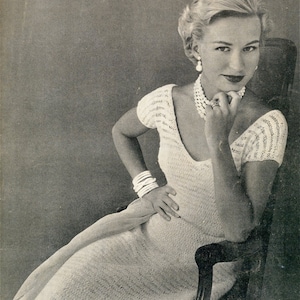 Può includere: Una donna indossa un abito bianco all'uncinetto con scollo rotondo e maniche corte. È seduta su una sedia e indossa una collana di perle e orecchini.
