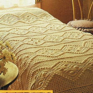 May include: A cream-colored, intricately crocheted bedspread with a detailed geometric pattern. The design features repeating diamond and wave-like motifs, creating a textured surface. The bedspread is draped over a bed, with a wooden chair visible in the background.