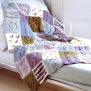 May include: A knitted patchwork throw blanket in shades of purple, blue, green, and white with a cable knit pattern. The blanket is draped over a light grey sofa.